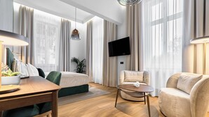 Ett modernt hotellrum med en säng, två fåtöljer, ett litet bord, en TV och stora fönster med gardiner.