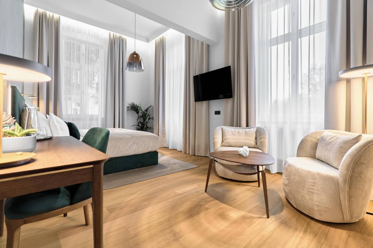 A modern hotel room with a bed, two armchairs, a small table, a TV, and large windows with curtains.