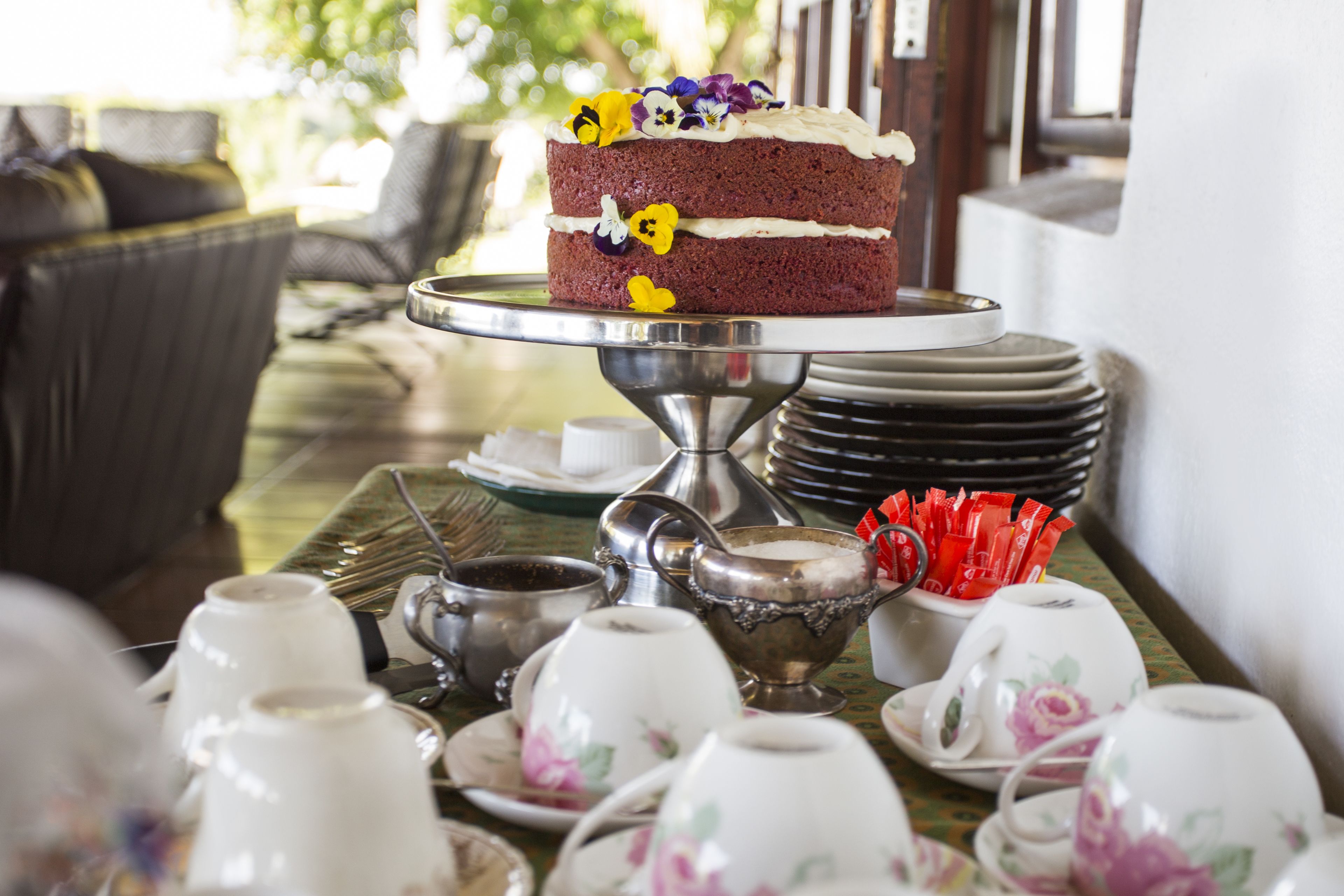 A table with a tiered cake stand holding a two-layered cake with floral decorations, surrounded by tea cups and saucers.