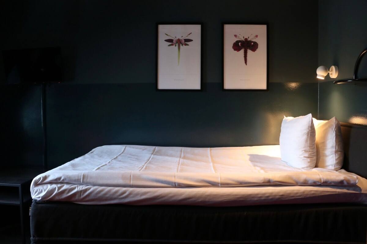 A neatly made bed with white linens and pillows, a dark wall, and two framed botanical illustrations above it.