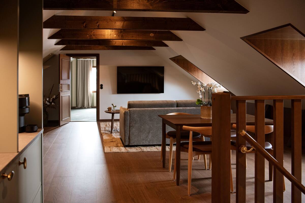 A modern interior with a dining area, wooden ceiling beams, a sofa, and a view into another room.