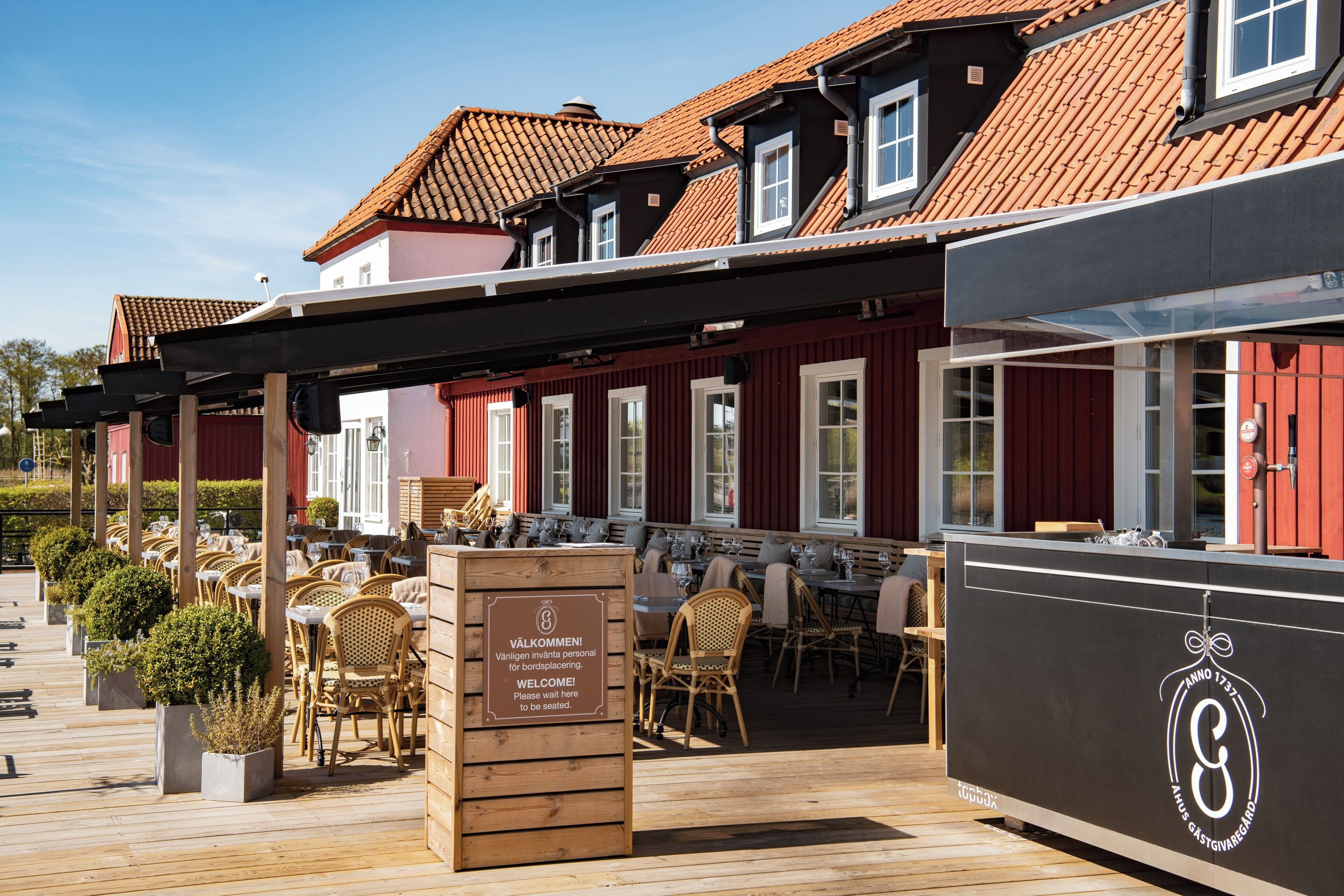 An outdoor seating area with wooden tables and chairs, a signboard, and a building with red walls and a tiled roof.