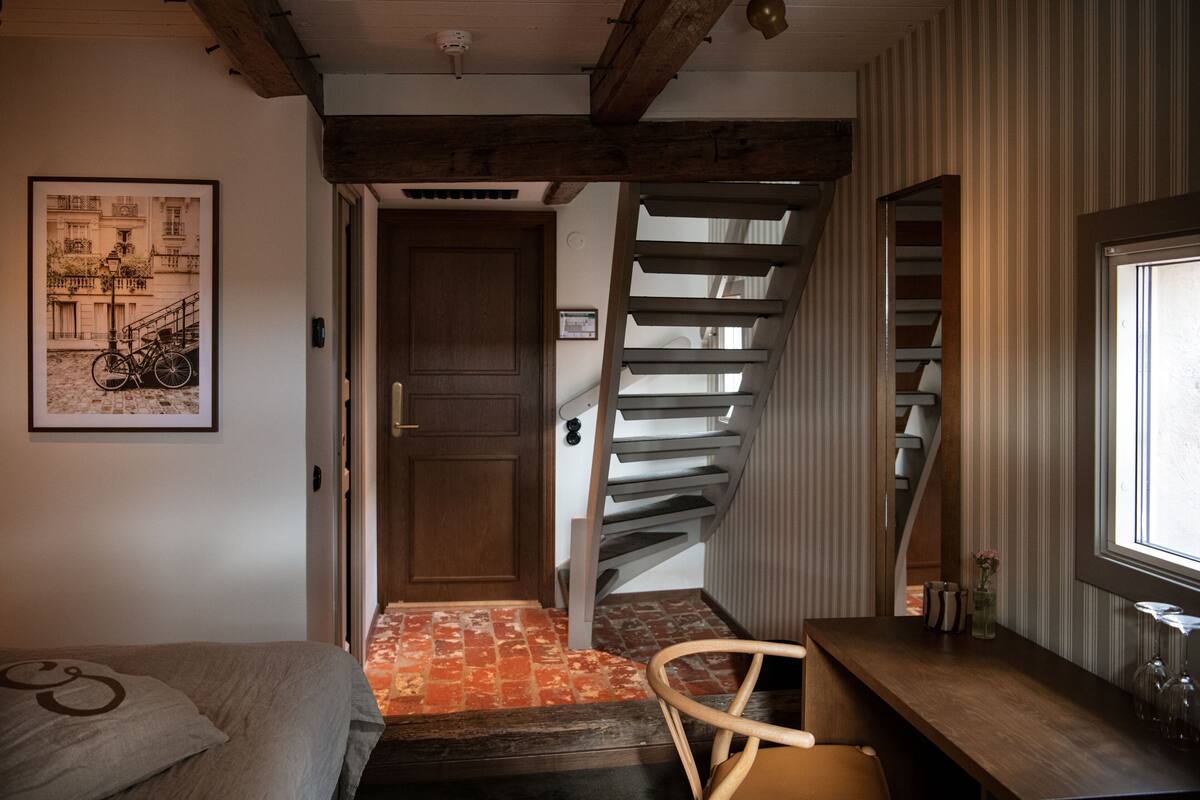 A room with a bed, a desk, a chair, a staircase, and a door leading to another room.