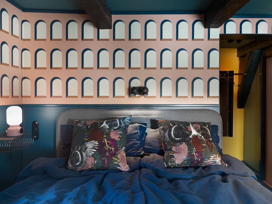 A bedroom with a bed, pillows, a wall-mounted lamp, and a ceiling with arched windows.