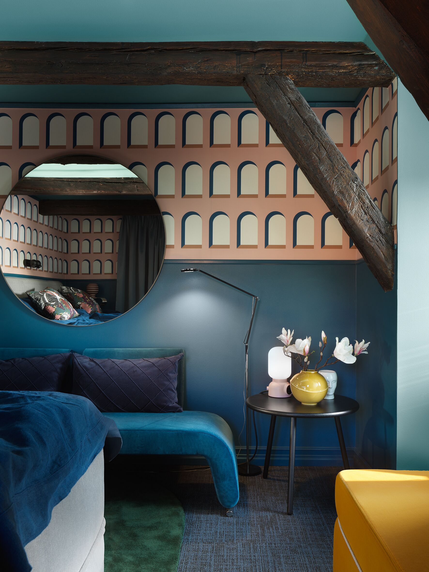 Une chambre avec un canapé bleu, un fauteuil jaune, un miroir rond et un mur orné d’un motif géométrique.