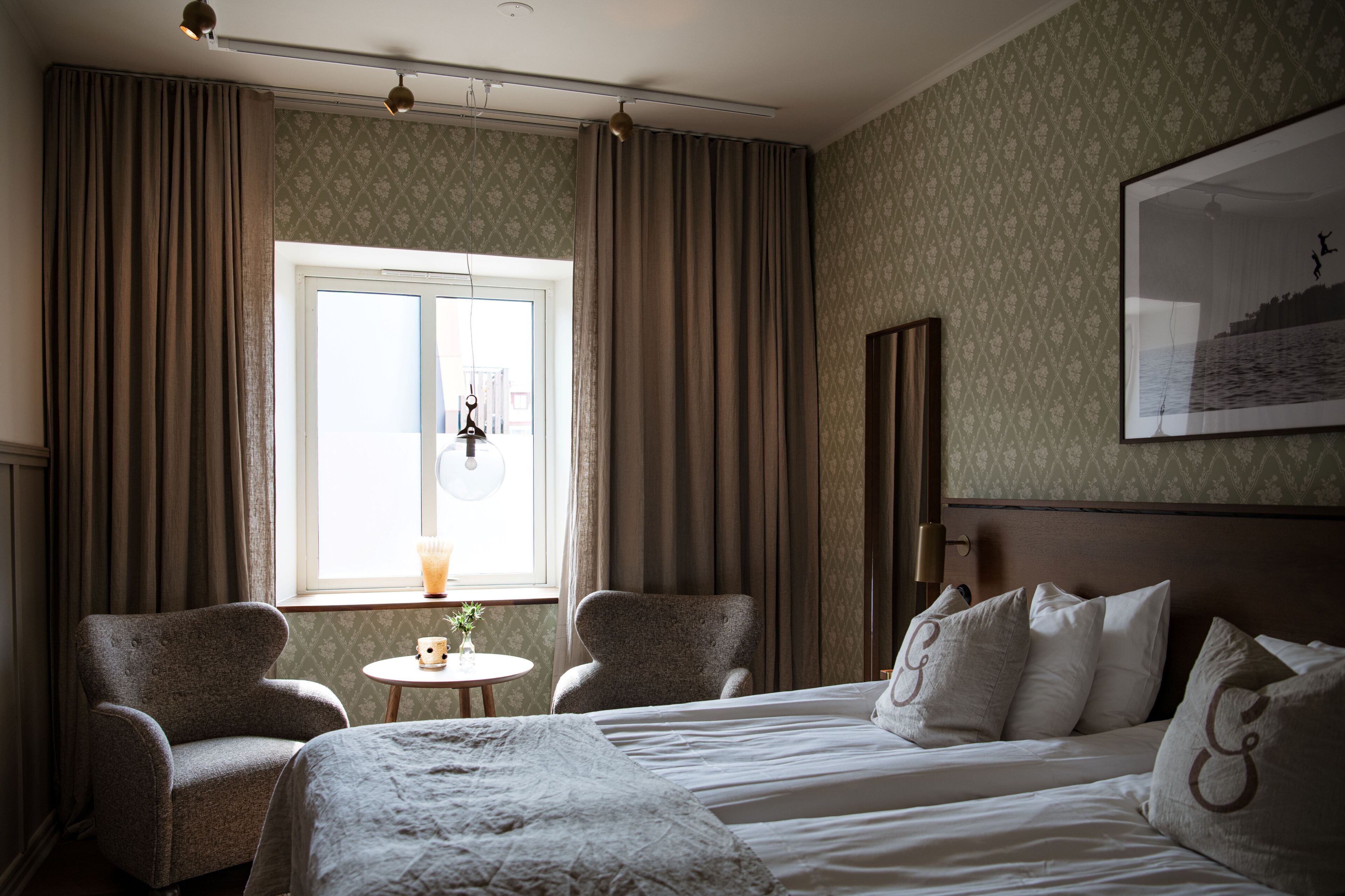 Habitación de hotel con cama, dos sillones, una mesita con una planta, ventana con cortinas e imagen en el marco en la pared.