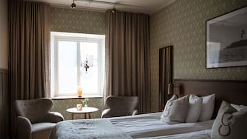 A hotel room with a bed, two armchairs, a small table with a plant, a window with curtains, and a framed picture on the wall.