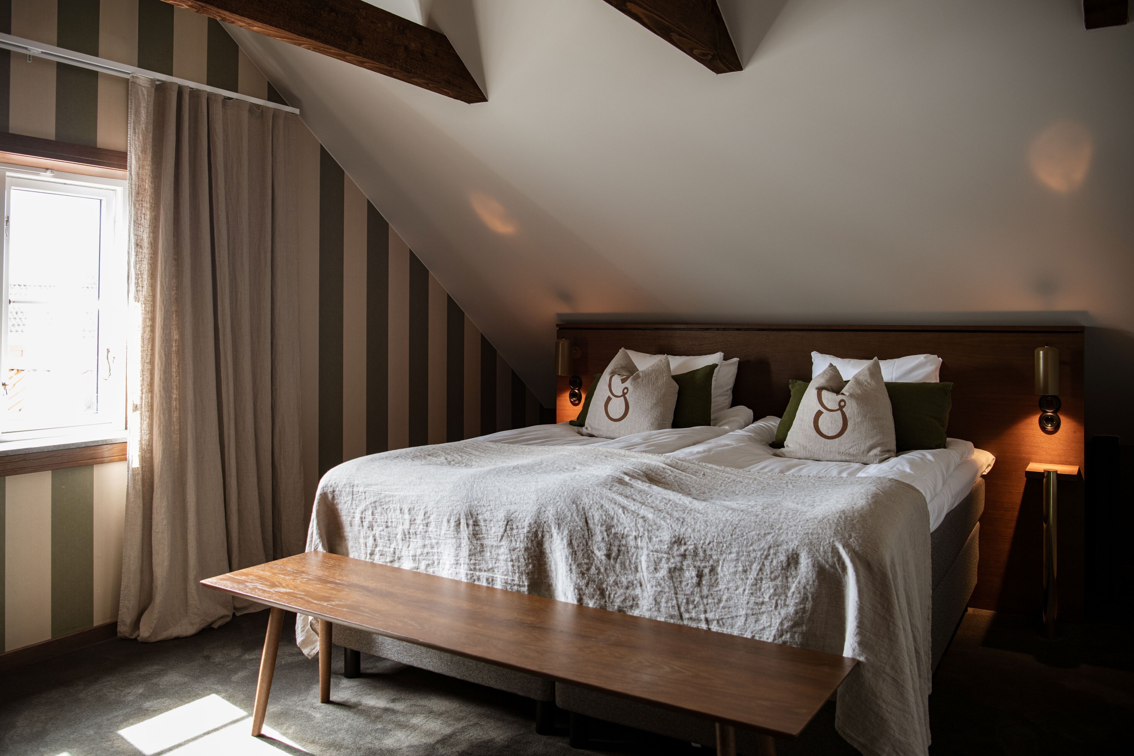 A bedroom with a bed, bedside lamps, a bench, a window with curtains, and striped walls.