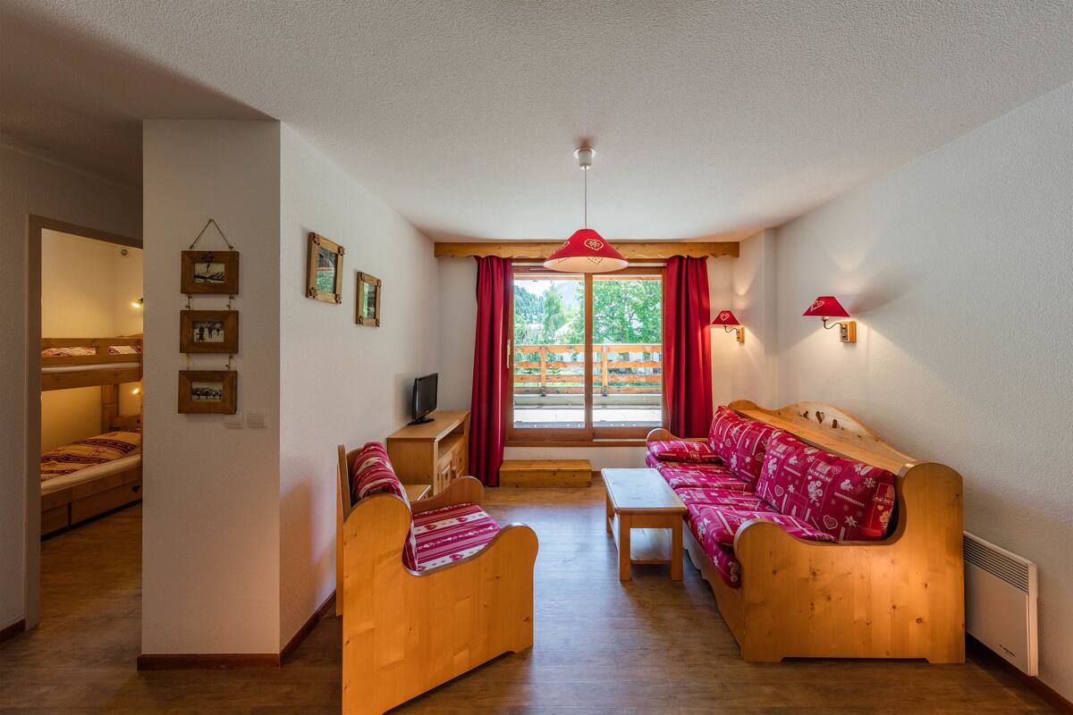 Una habitación acogedora con un sofá de madera, una mesita y un televisor. Hay cortinas rojas y una ventana con vistas a árboles.