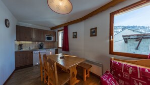A cozy kitchen with wooden cabinets, a dining table, and chairs. There is a window with a view of a snowy landscape. - Noemys Les Balcons du Viso  (Abriès-Ristolas)
