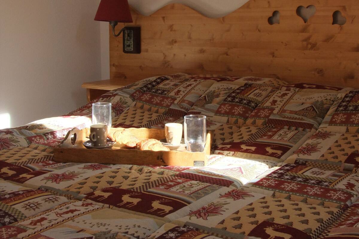 Una cama con cabecera de madera, colcha con estampados de animales, una bandeja con taza y vaso, y una lámpara roja sobre una mesita de noche.