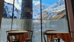 A room with a large window offering a view of snow-covered mountains. There are two tables with chairs, and a stone pillar is visible in the corner. - Hotel Aigle (Courmayeur)