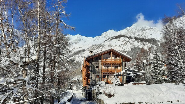 A snow-covered mountain lodge with a wooden exterior and a balcony, surrounded by snow-laden trees and a clear blue sky. - Hotel Aigle (Courmayeur)