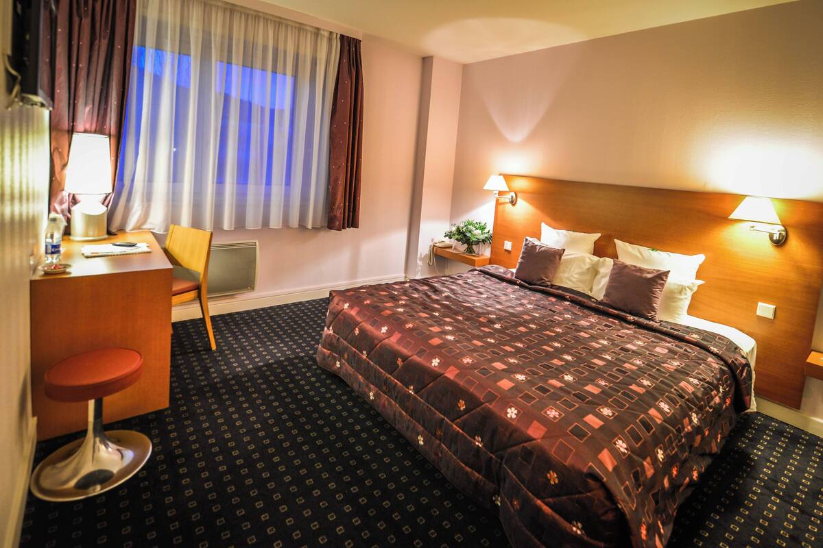 Una habitación de hotel con una cama grande, un escritorio con una silla, un taburete pequeño, una mesita de noche con una lámpara y una ventana con cortinas.