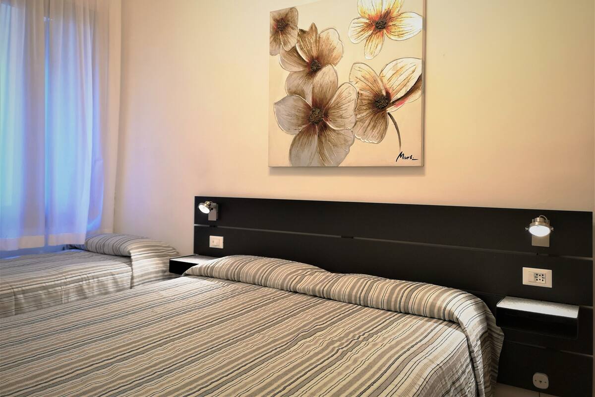 Un dormitorio con cama, mesita de noche, lámpara de pared y un cuadro de flores.