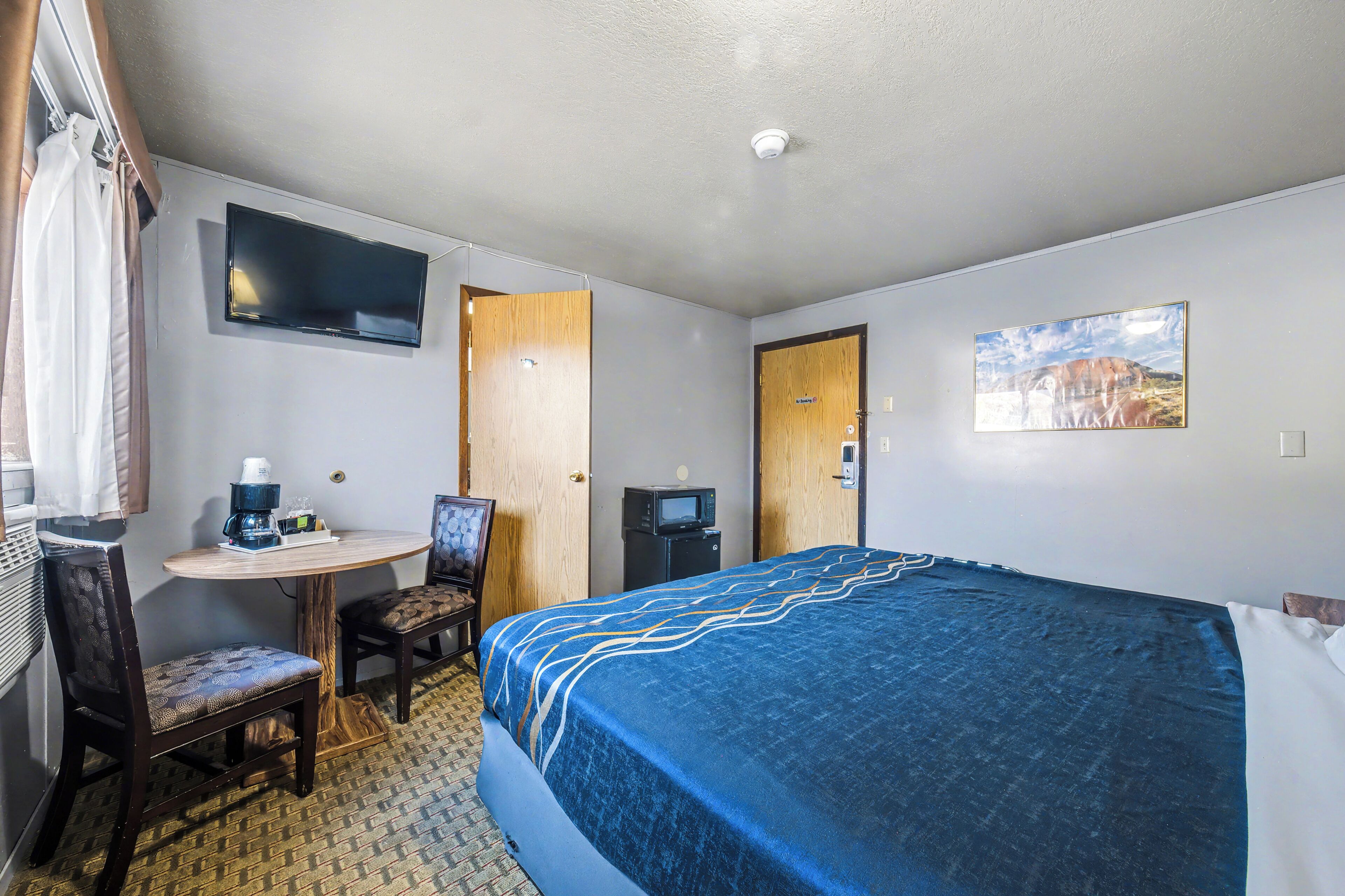 A hotel room with a bed, a desk with a chair, a television mounted on the wall, and a door.