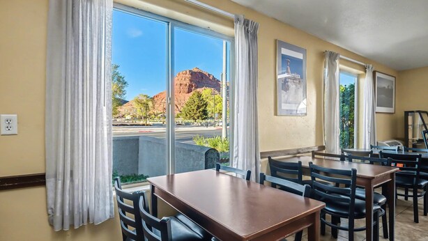 Habitación de hotel con ventanas amplias, un comedor con mesas y sillas, y vistas a una montaña.