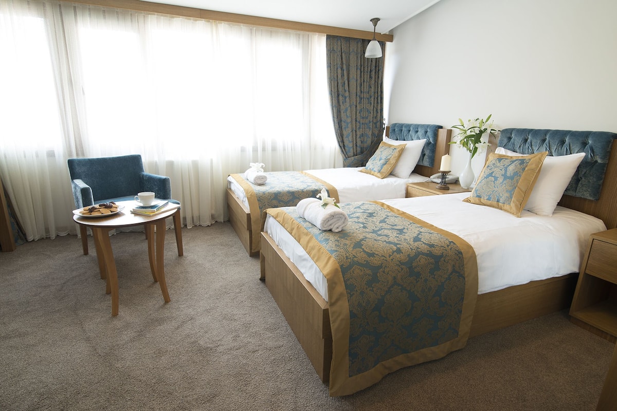 Een hotelkamer met twee bedden, een klein tafeltje met een stoel en een raam met gordijnen.
