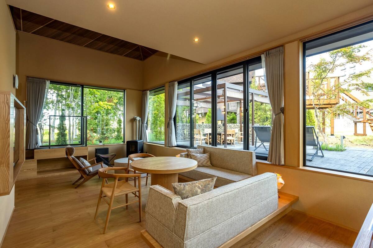 Amplia sala de estar con sofá, sillas y una mesa redonda. Grandes ventanales ofrecen vistas a muebles de exterior y follaje.