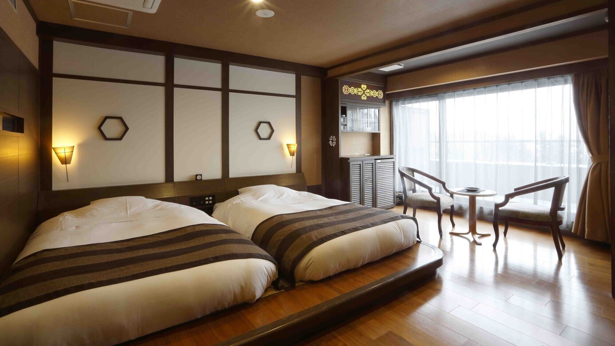 A hotel room with two beds, a small table with chairs, a minibar, and a large window with curtains.