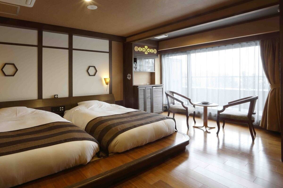 Habitación de hotel con dos camas, una pequeña mesa con sillas, un minibar y un ventanal con cortinas.