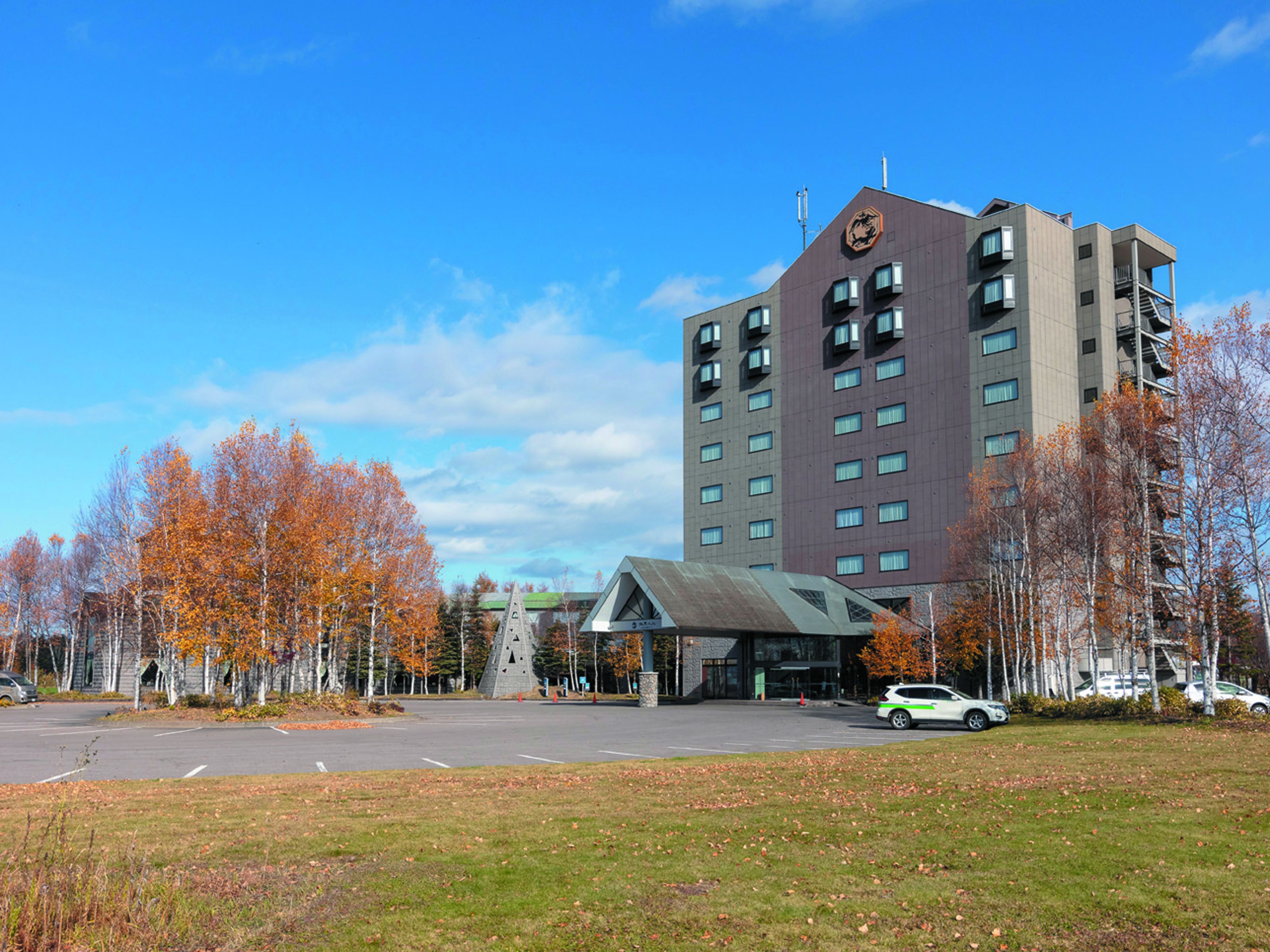 A modern hotel with a central entrance, surrounded by autumn trees and a parking lot.