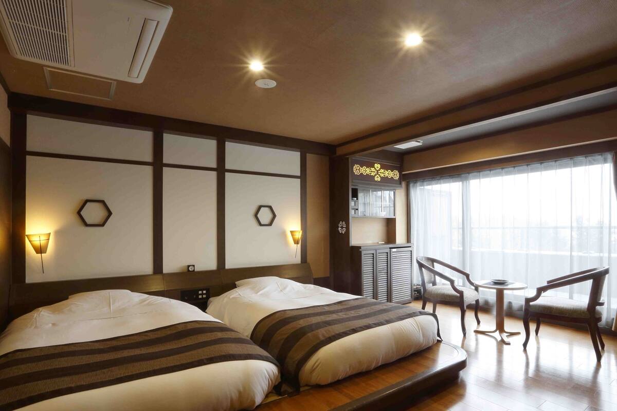 Habitación de hotel con dos camas, un escritorio, sillas, un ventanal grande y un ventilador de techo.