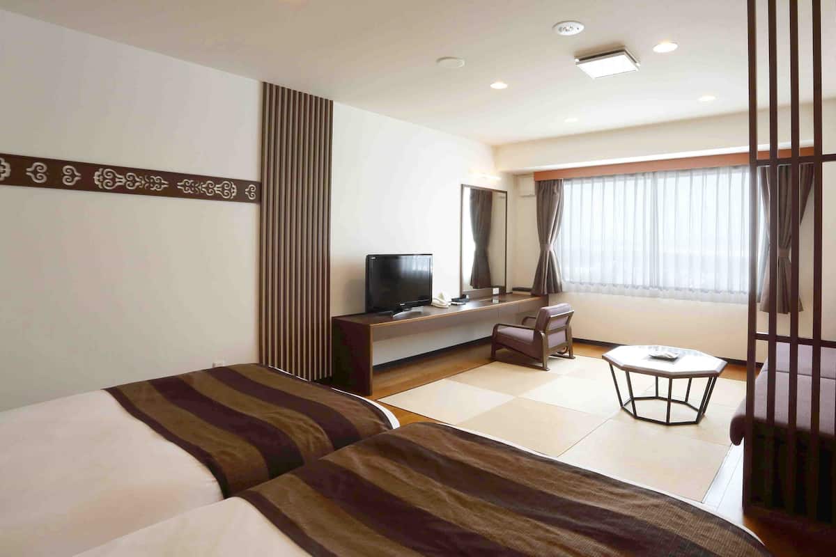 Una habitación de hotel con cama, escritorio con televisor, silla, mesita y ventana con cortinas.