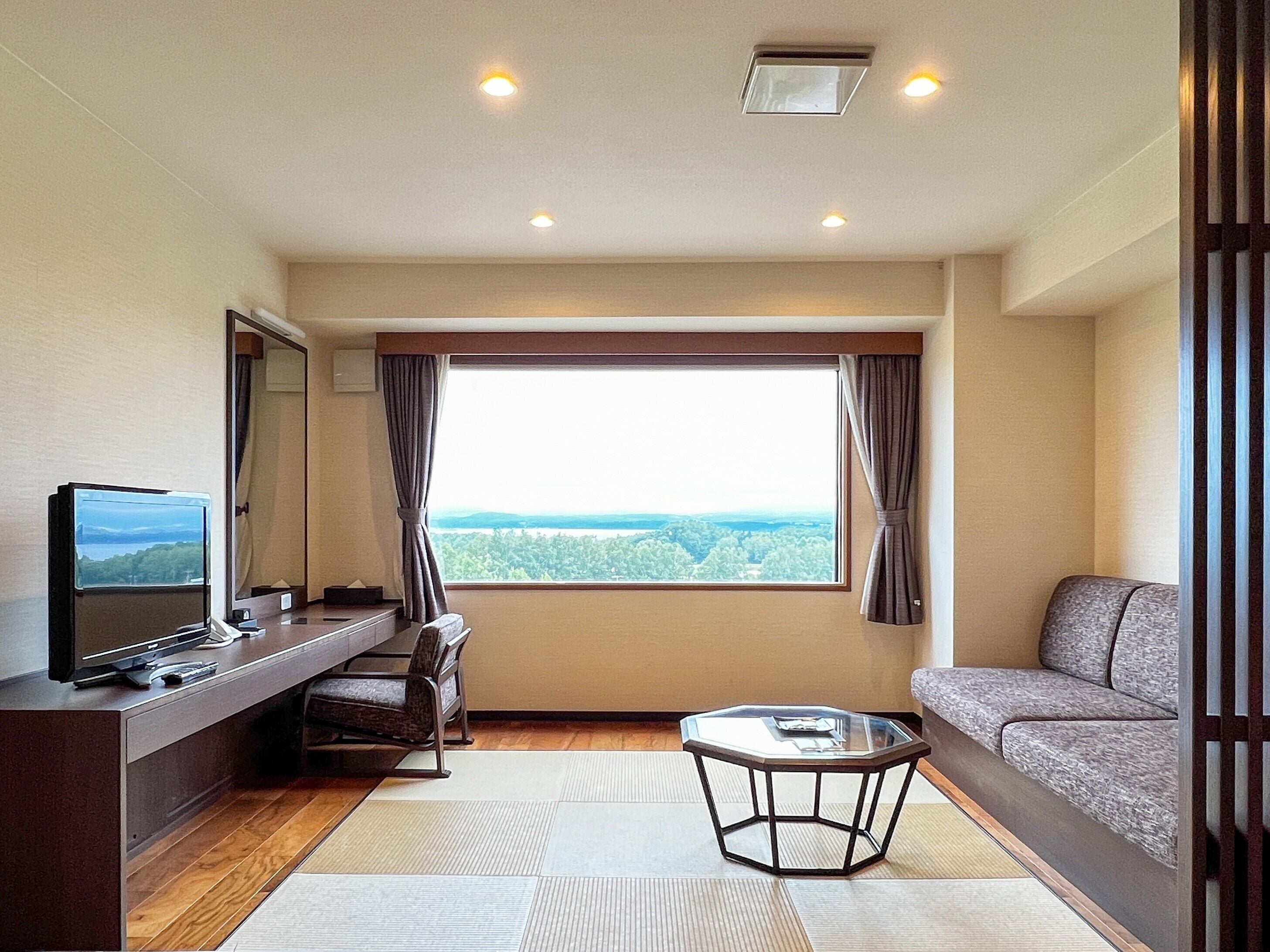 A hotel room with a large window, a flat-screen TV, a sofa, a small table, and a desk with a computer.