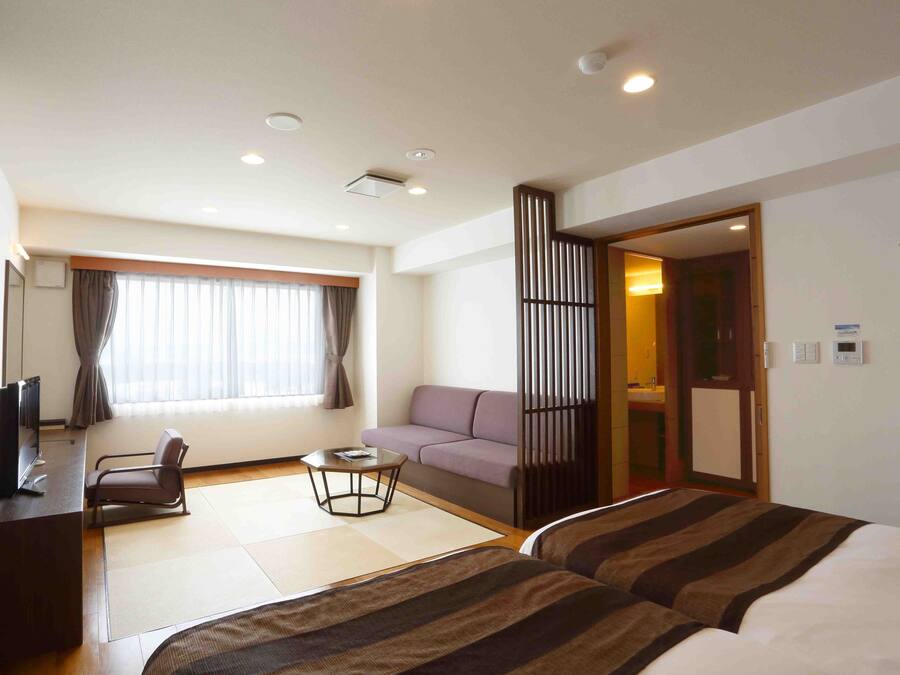 A hotel room with a bed, a sofa, a desk with a chair, a small table, and a window with curtains.