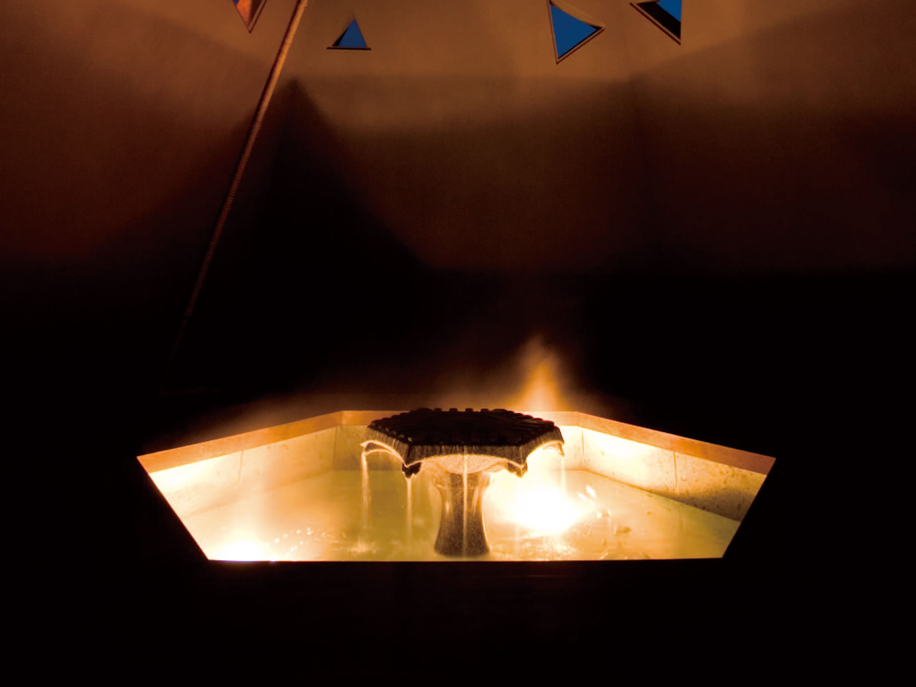 Una habitación con poca luz, una mesa en el centro y una silla, un respiradero en el techo y una lámpara fijada a la pared.