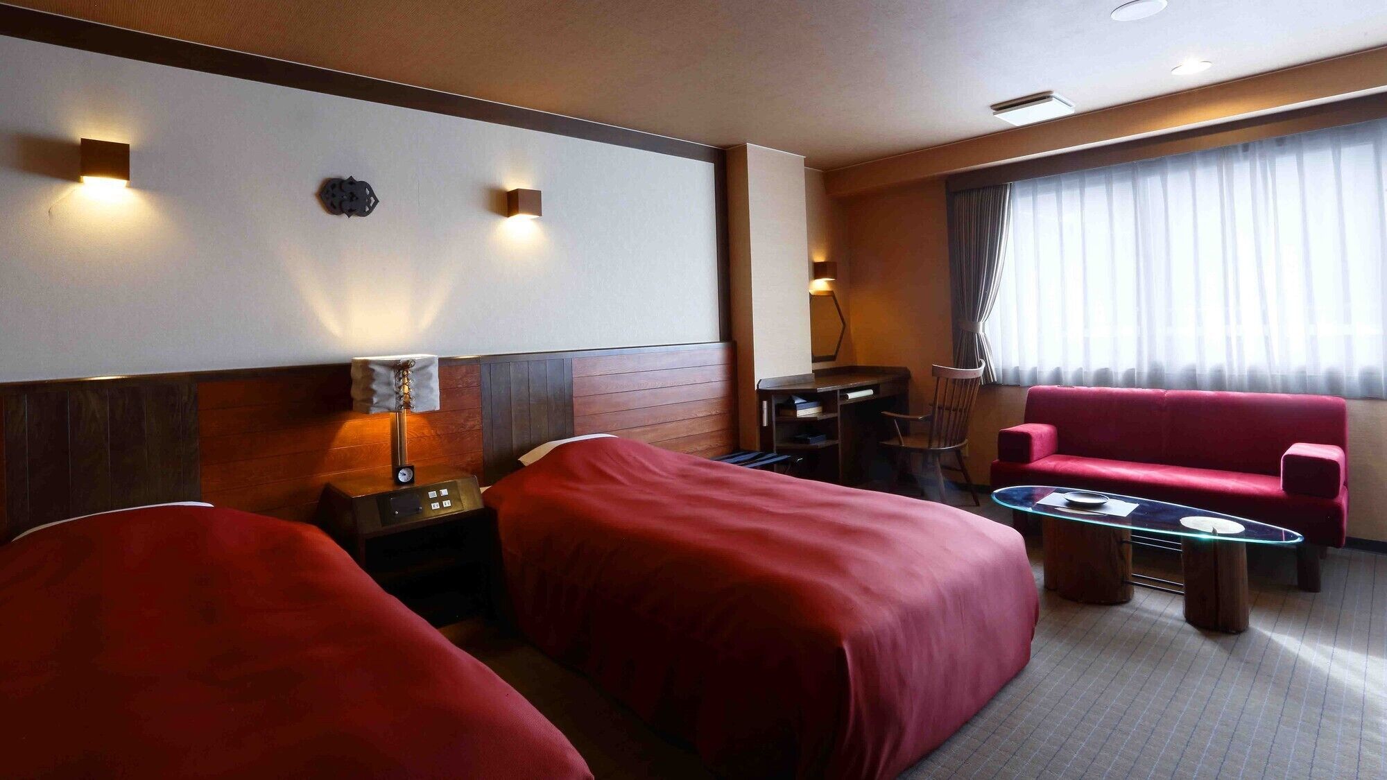 A hotel room with two beds, a desk, a chair, a red sofa, and a window with curtains.
