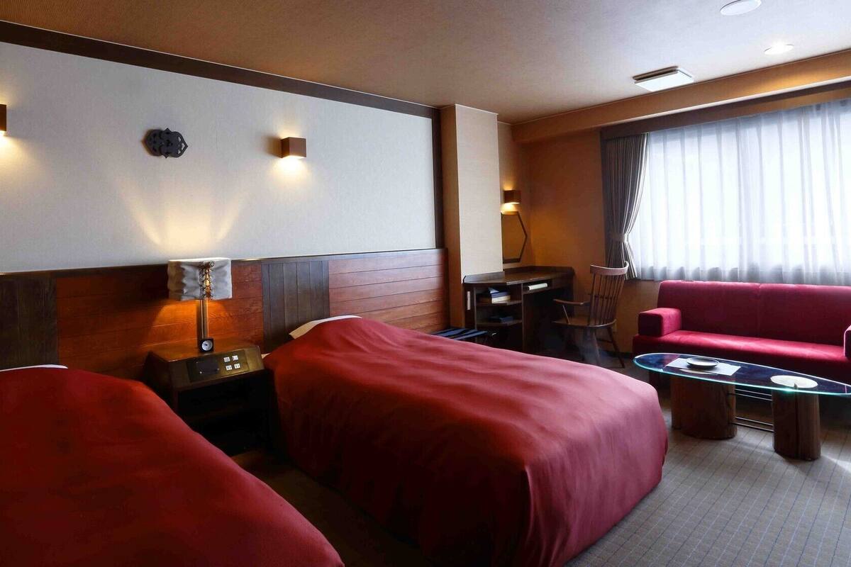 Habitación de hotel con dos camas, un escritorio, una silla, un sofá rojo y un ventanal con cortinas.