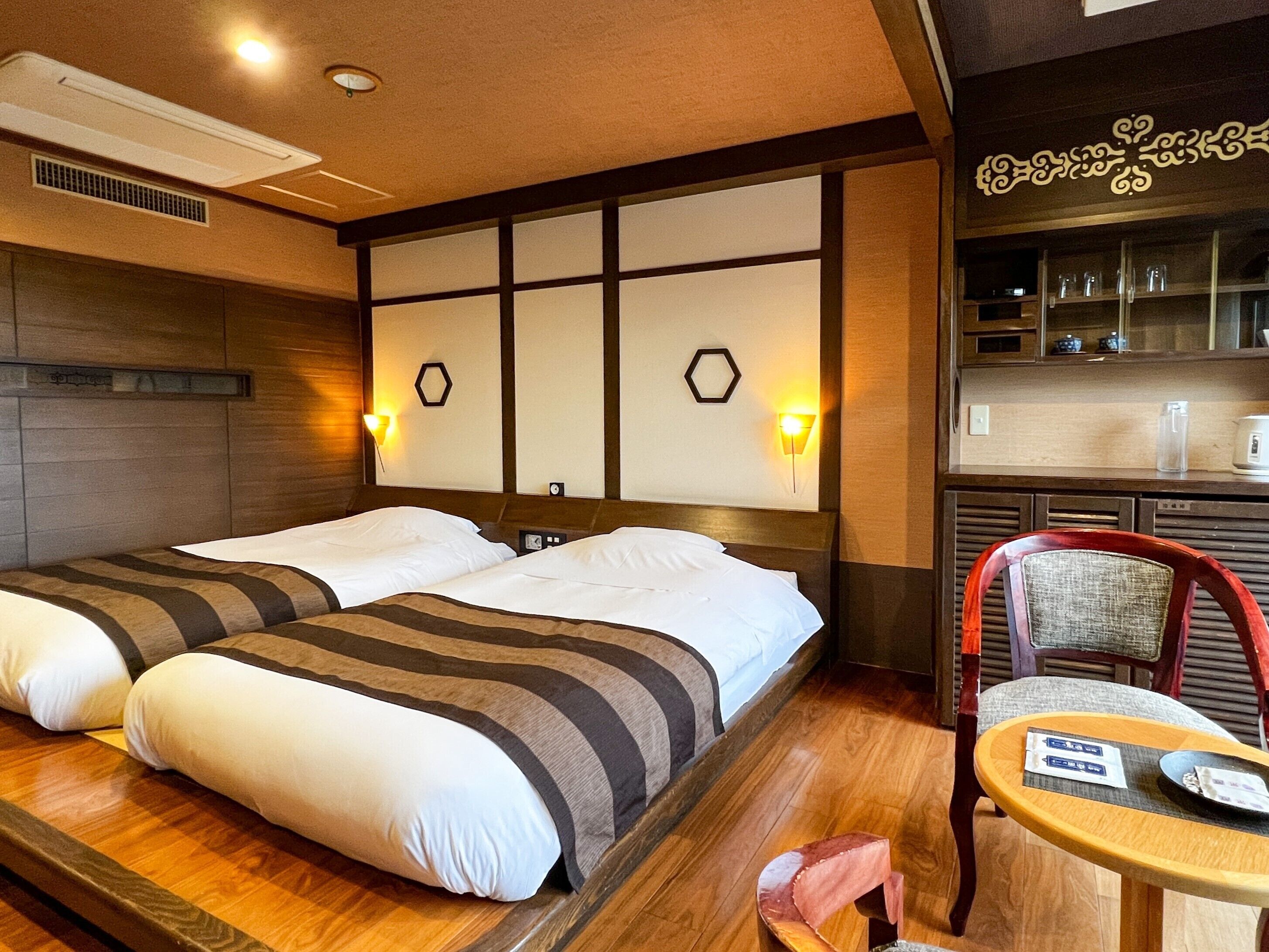 Habitación de hotel con dos camas, suelo de madera, una mesita con una silla y una zona de cocina con estanterías.