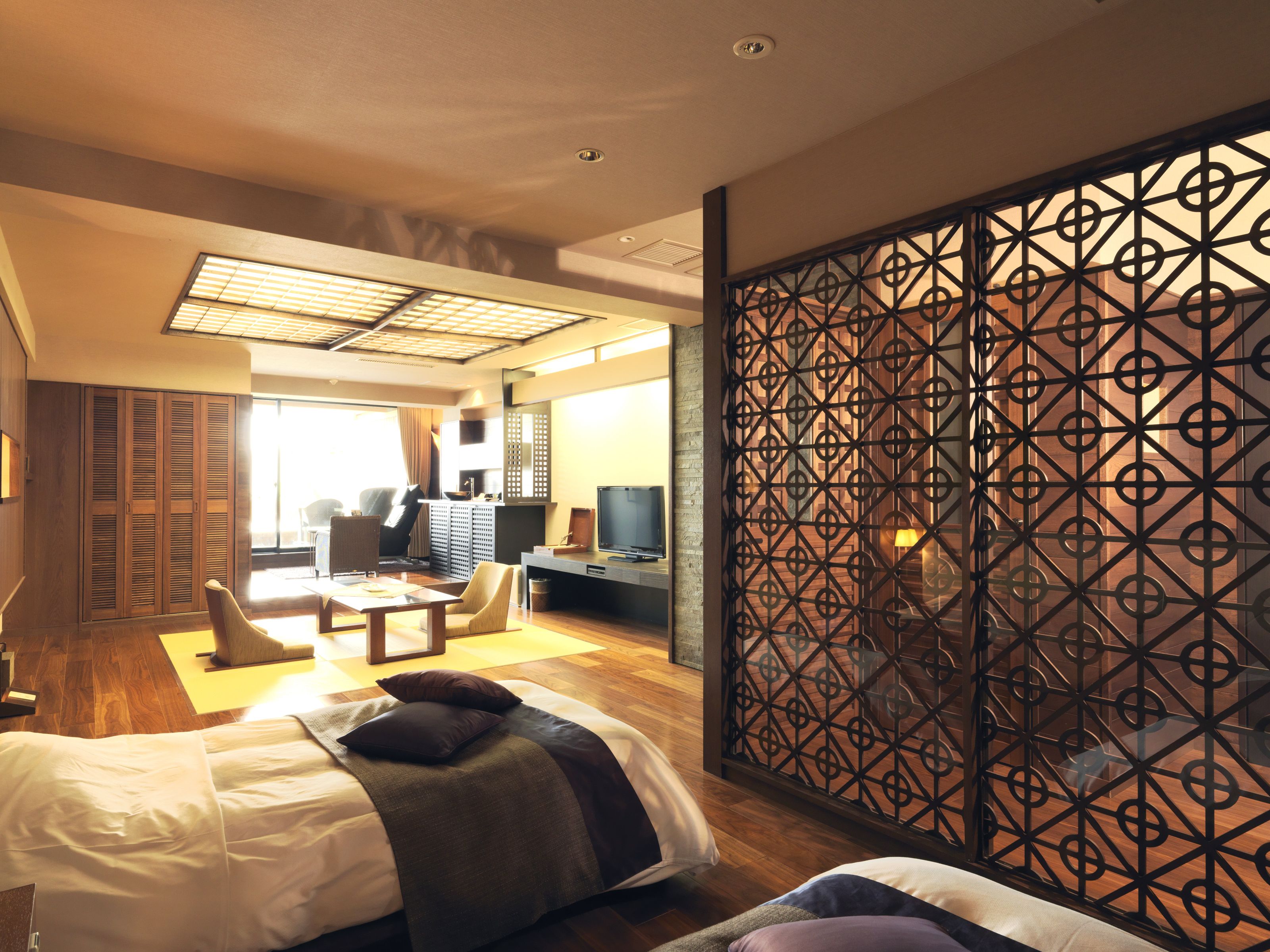 A hotel room with a bed, a desk, a chair, a TV, and a partition with a geometric pattern.