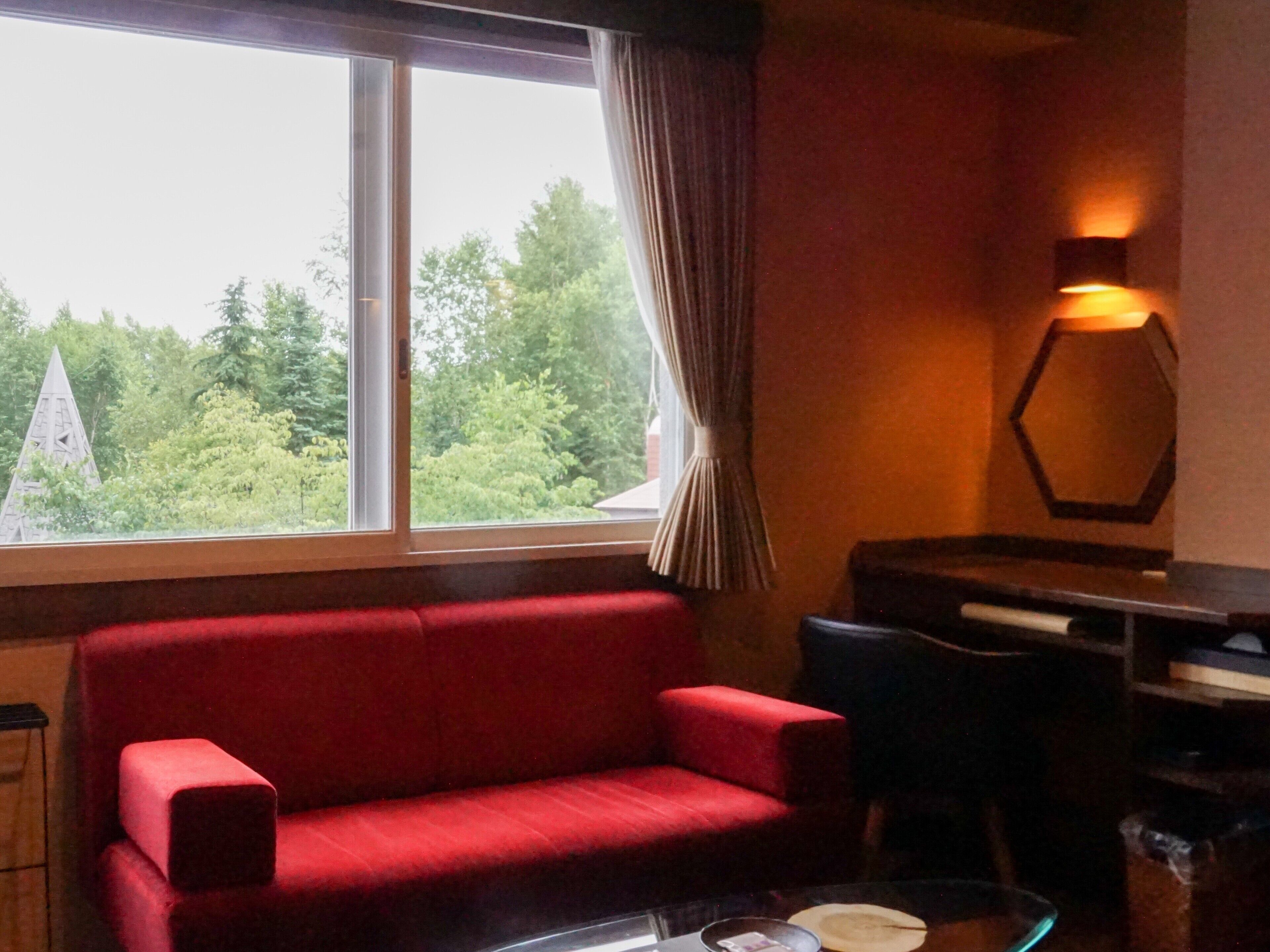 Una habitación con un sofá rojo, una mesa de centro de cristal, una silla y vistas a árboles y un edificio a través de la ventana.
