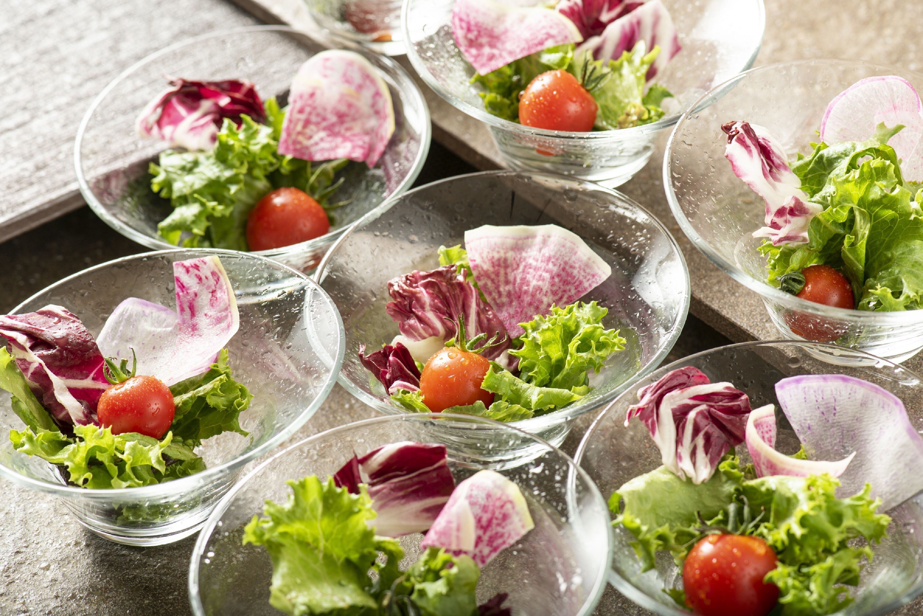 A series of clear glass bowls containing fresh salads with lettuce, radishes, and cherry tomatoes.