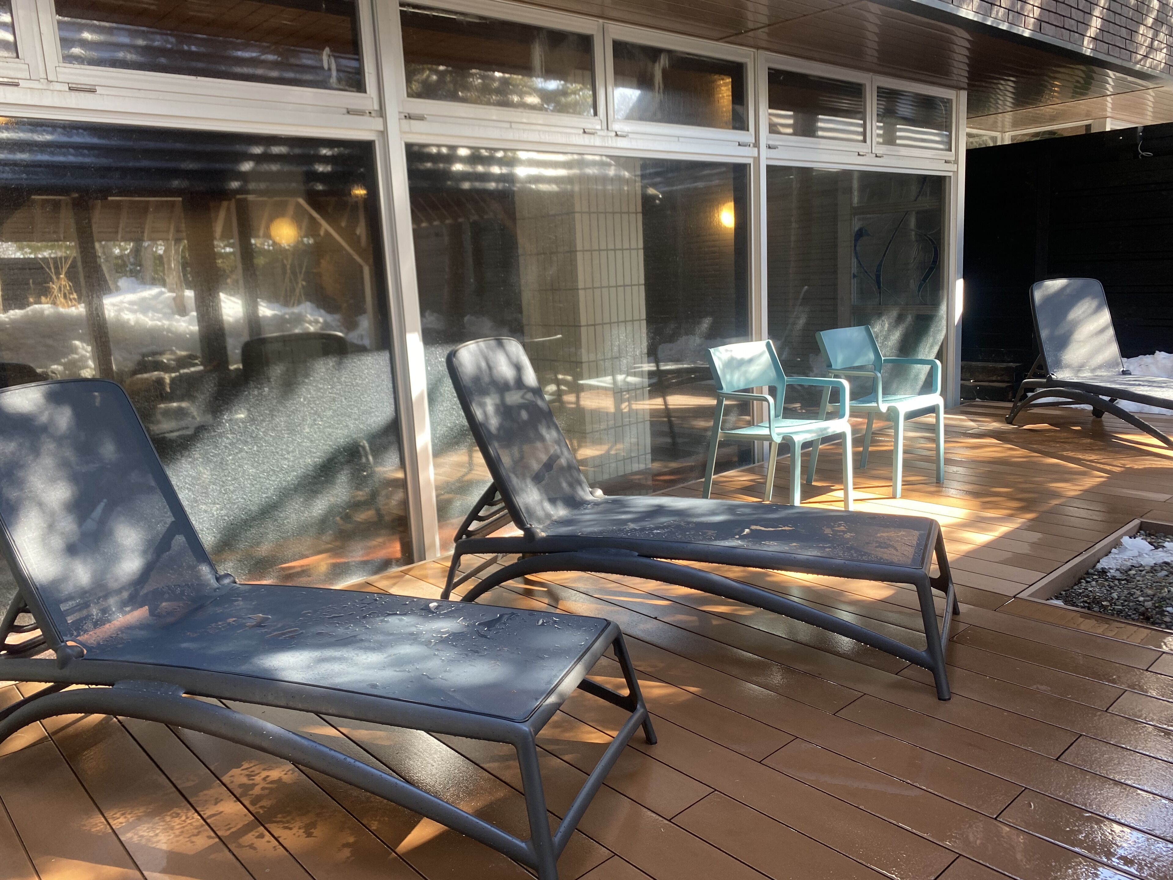 A covered patio with two lounge chairs and a table, large windows, and a view of a snowy landscape.