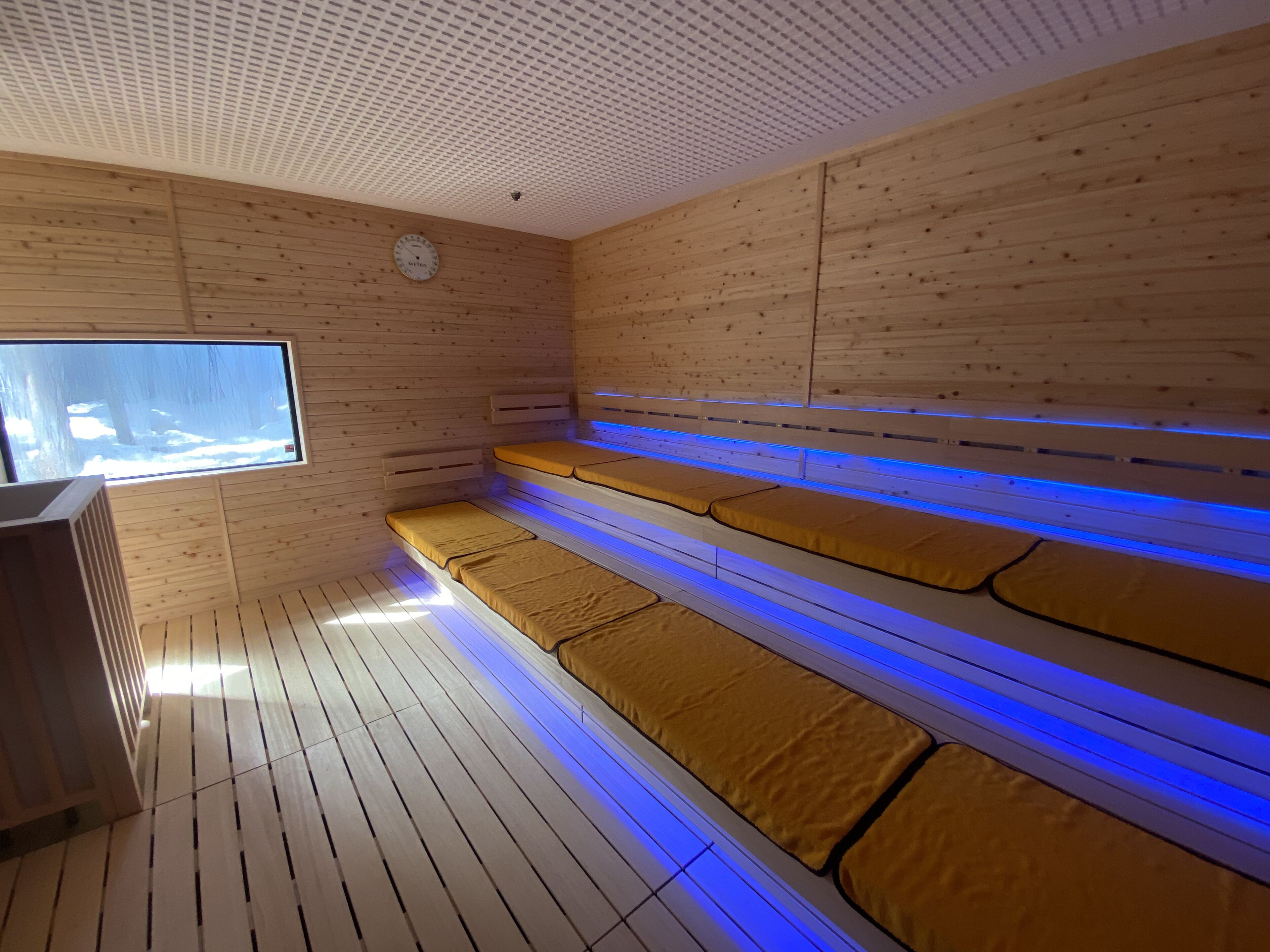 Una sala de sauna con paredes y bancos de madera, un ventanal grande y un techo con diseño perforado.