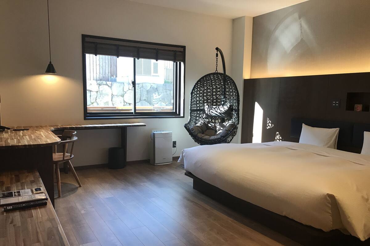 Una habitación de hotel moderna con suelo de madera, una cama con ropa blanca, una silla colgante, un escritorio con silla y un gran ventanal con persianas.