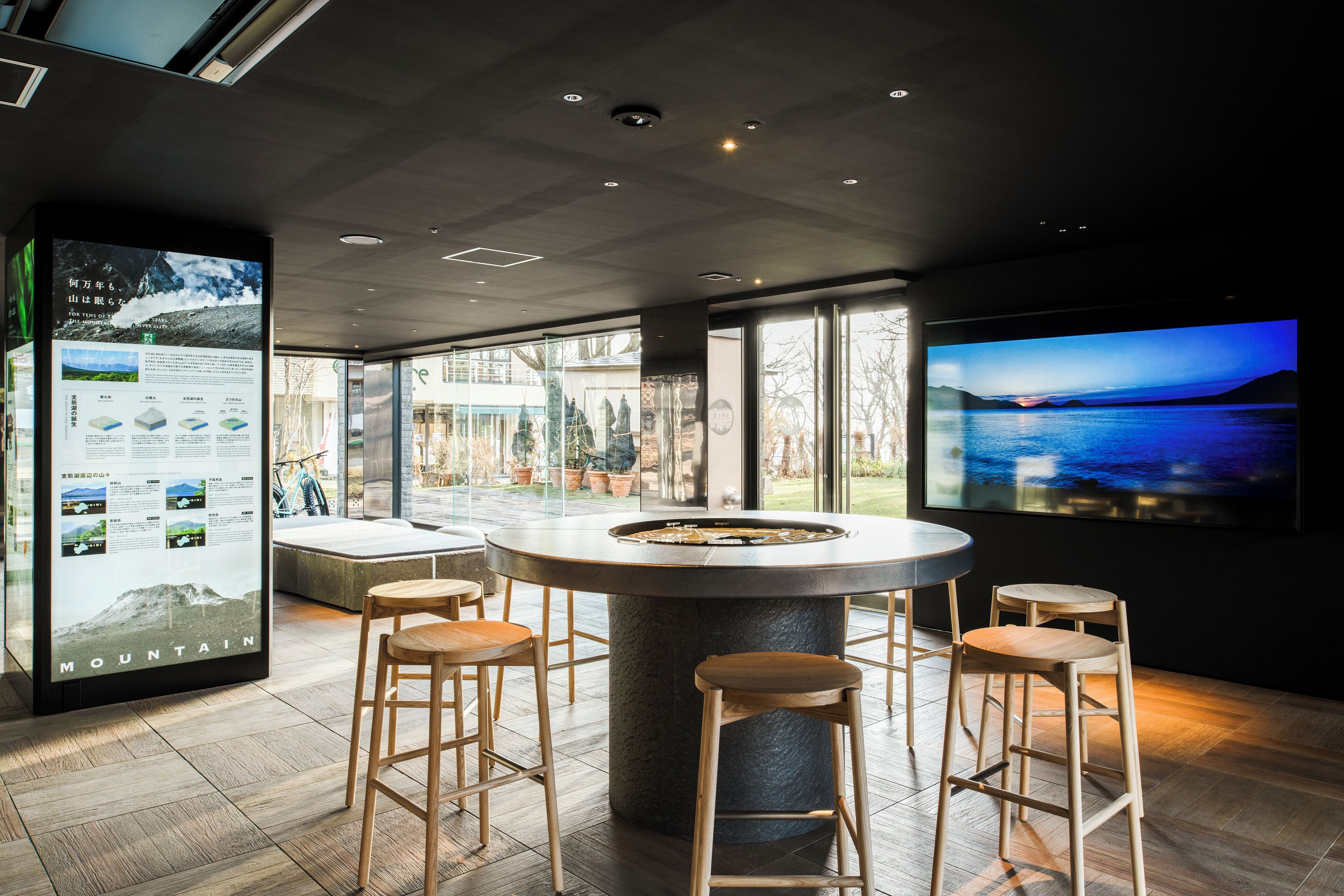 A modern bar area with a round table, wooden stools, and a large screen displaying a scenic view.