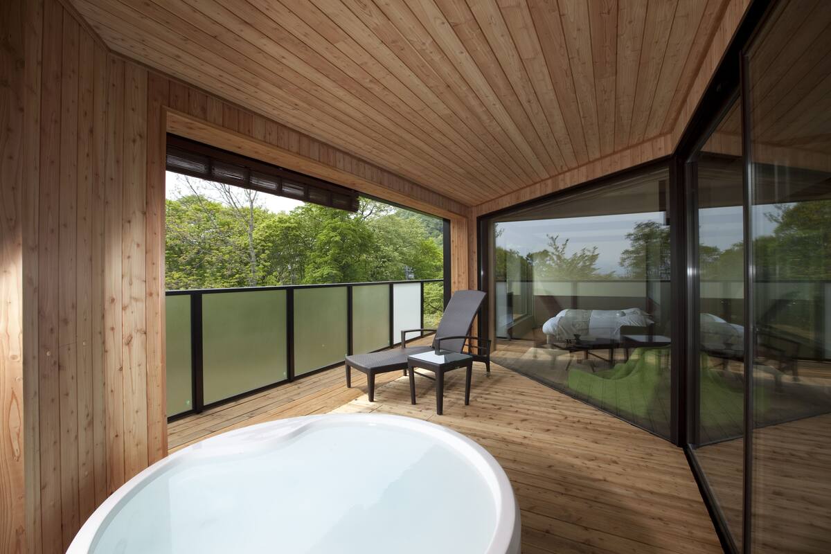 Una terraza de madera con un jacuzzi, dos sillas y un balcón acristalado que da a un paisaje verde.