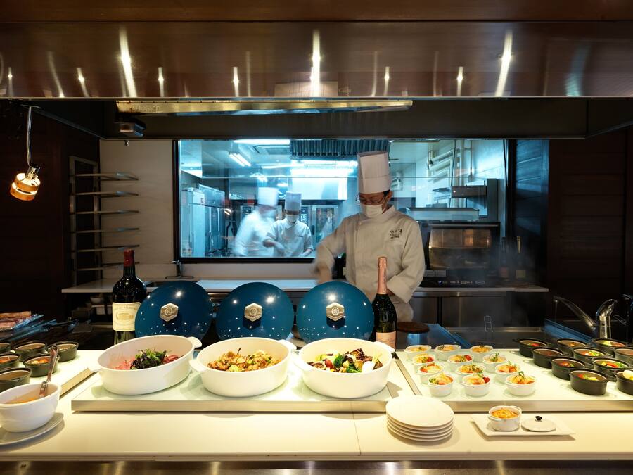 Un chef prepara alimentos en una cocina moderna con un ventanal grande, diferentes estaciones de cocina y un bufé con varios platos.