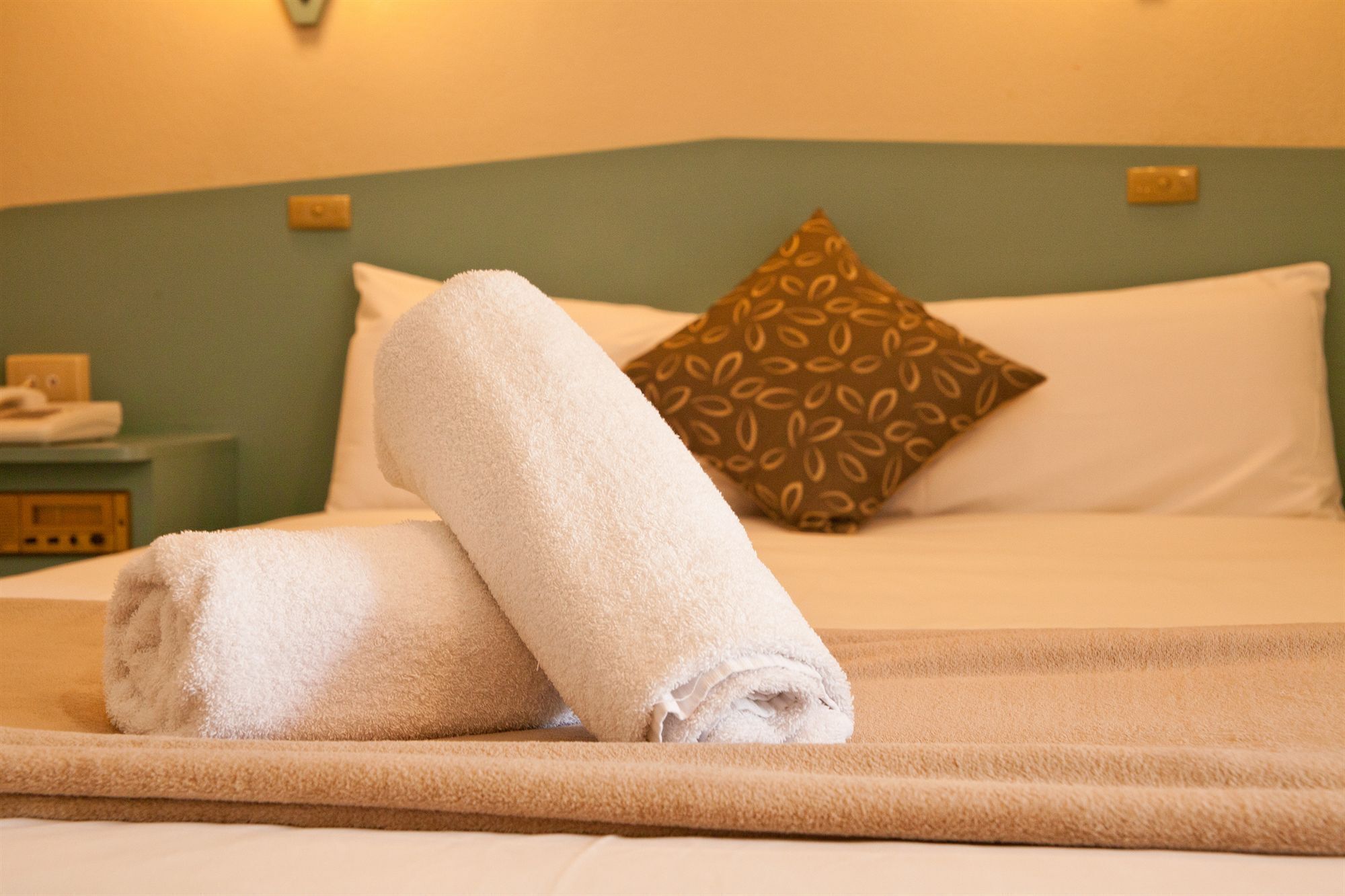 A neatly made bed with a rolled white towel and a brown patterned pillow.