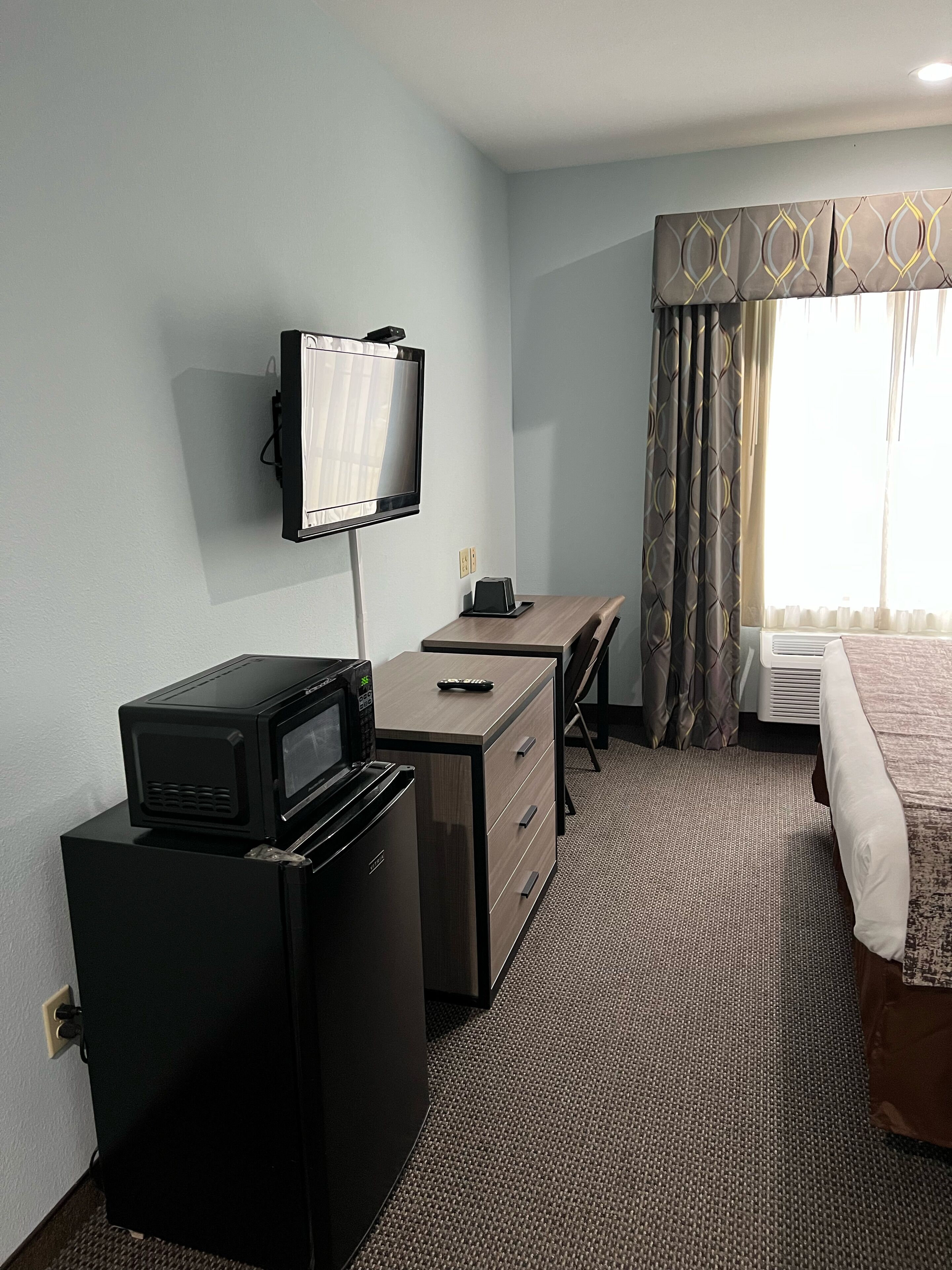 Quarto de hotel com televisão de tela plana na parede, um pequeno refrigerador preto, uma mesa com cadeira, uma cama com colcha estampada e uma janela com cortinas.