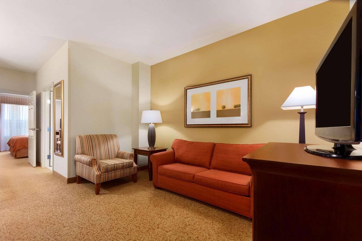 A hotel room with a sofa, armchair, TV, and framed artwork on the wall.