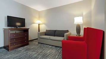 A hotel room with a flat-screen TV on a wooden stand, a beige sofa, a red armchair, a floor lamp, and a lamp with a striped shade.