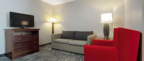 Habitación de hotel con televisor de pantalla plana sobre un soporte de madera, un sofá color beige, un sillón rojo, una lámpara de pie y una lámpara con pantalla a rayas.