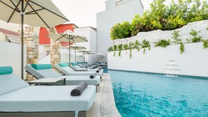 A poolside area with lounge chairs and umbrellas, a stone wall, and a white building with greenery.