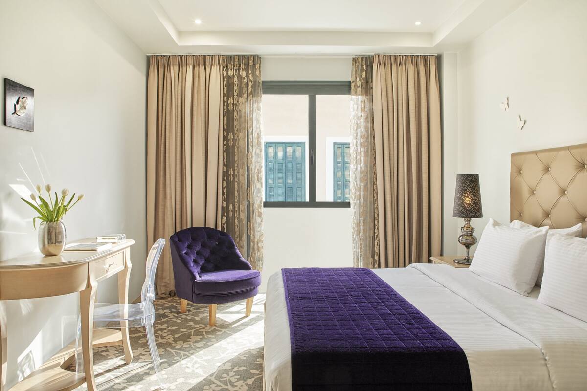 A hotel room with a bed, a desk, a chair, a window with curtains, and a lamp.